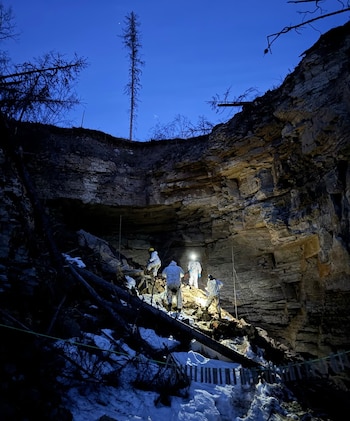 Cuatro personas con trajes protectores y linternas frontales en una ladera rocosa. Hay troncos de árboles caídos, nieve y un árbol seco bajo cielo azul nocturno
