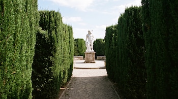 Escultura blanca de un minotauro sobre un pedestal de piedra al final de un camino flanqueado por altos setos verdes de ciprés.