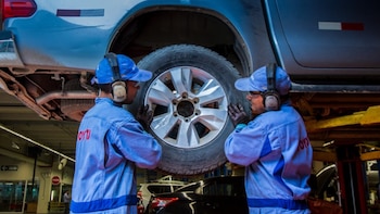 Dos mecánicos con uniforme azul y protección auditiva revisan una rueda de una camioneta elevada en un taller mecánico