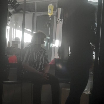 Una bolsa de suero amarillo colgando, con un tubo conectándose. Se ven varias personas borrosas sentadas en una habitación iluminada por ventanas