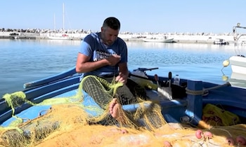 Hombre descalzo sentado en una embarcación azul. Repara redes de pesca amarillas. Fondo con agua, muelle de rocas y barcos