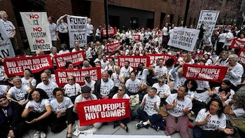 Multitud de personas protestando en una calle urbana. Muchos llevan camisetas blancas y sostienen pancartas rojas y blancas con lemas anti-guerra y pro-Palestina