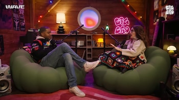 Dos personas sentadas en sillones tipo Beanbag Chair verdes frente a micrófonos en un estudio con paredes de madera. La mujer tiene una manta tipo Cushion colorida sobre las piernas
