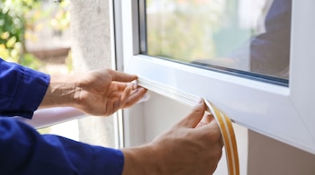 Primer plano de unas manos aplicando una tira de burlete adhesivo de color blanco y beige en el marco de una ventana de PVC, con luz natural de fondo.