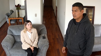 Mujer joven con micrófono sentada en sofá gris, hombre con sudadera gris de pie, mesa de comedor con flores y sillas, suelo de madera, paredes blancas.