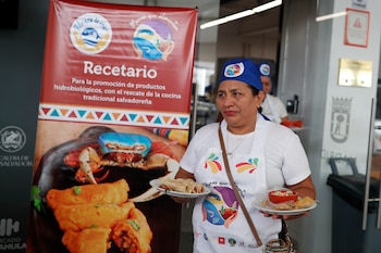 Una mujer sonriente con gorro y delantal blanco con un logo de comida, sostiene una bandeja negra con tres platos de comida. Al fondo, personas y pancartas