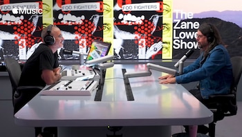 Dave Grohl y Zane Lowe sentados en una mesa gris de estudio con micrófonos, el fondo muestra carteles de Foo Fighters y un paisaje de montaña