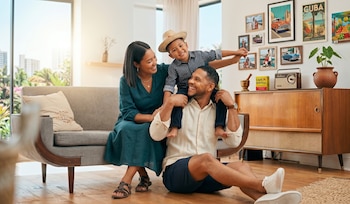 Un hombre, una mujer y un niño sonríen en una sala. El niño está en los hombros del hombre, la mujer sentada en un sofá gris. Hay cuadros cubanos y una radio