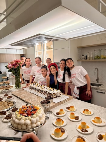 Un grupo de mujeres posa sonriente en una cocina detrás de una mesa servida con tartas, flanes y un florero