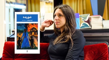 Una mujer sentada en un sillón rojo junto a la portada de un libro con texto en persa y una ilustración de una casa incendiada.