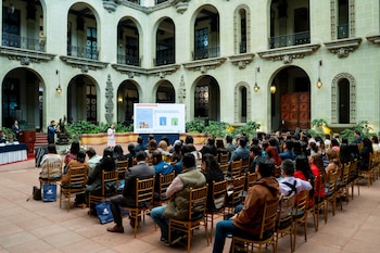 Un hombre de traje habla en un atril de madera frente a banderas institucionales, junto a un intérprete de lengua de señas