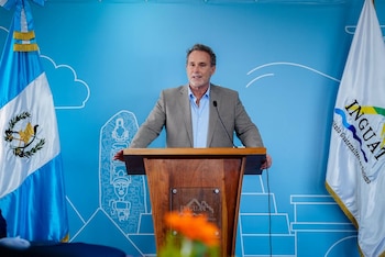 Un hombre de pie detrás de un podio de madera con el logo de INGUAT, con banderas de Guatemala e INGUAT y una pared azul con arte lineal al fondo