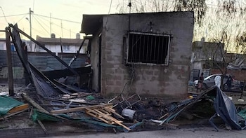 Edificio de concreto parcialmente destruido con ventana enrejada. Escombros, maderas quemadas, chapas metálicas y material verde en el suelo. Postes de luz y árboles al fondo