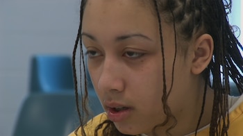 Primer plano del rostro de una mujer joven con trenzas largas. Viste una prenda de color amarillo o naranja. El fondo es un muro claro y difuminado