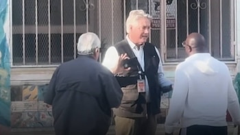 Un hombre con camisa blanca, chaleco oscuro y credencial habla con dos hombres en una calle urbana. Hay un edificio con ventanas enrejadas detrás