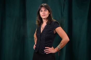 Lady M, mujer de cabello castaño, vestida de negro con joyas de plata y brazalete rojo, manos en las caderas, fondo de tela verde oscuro