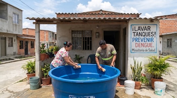 Una mujer y un hombre limpian un tanque de agua azul grande con cepillos en el exterior de una casa, junto a un cartel informativo.