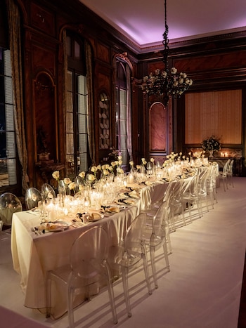 Mesa larga de cena preparada con mantel blanco, rosas blancas en jarrones, velas encendidas y sillas transparentes en un salón con paneles de madera oscura