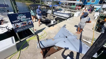 Un dron gris con forma de delta reposa sobre el suelo de concreto de un muelle junto a un cartel publicitario, barcos y personas