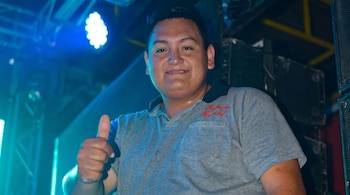 Un hombre con una camisa polo gris sonríe y levanta el pulgar, iluminado por una fuerte luz azul, en lo que parece ser un escenario con equipo oscuro