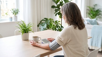 Mujer adulta sentada en mesa de comedor midiéndose la presión arterial con tensiómetro digital. Fondo de hogar luminoso con plantas y taza de té en la mesa.