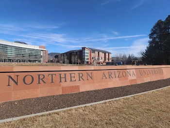 Un muro de piedra roja con el texto Northern Arizona University frente a edificios académicos y un cielo despejado