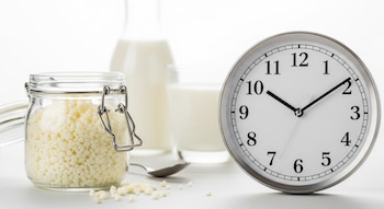 Un frasco de vidrio con gránulos de kéfir de leche en primer plano y un reloj analógico grande. Una cuchara, un vaso y una jarra de leche al fondo.