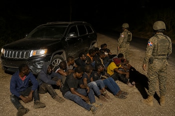 Varios hombres están sentados en el suelo junto a un automóvil negro con las luces encendidas, custodiados por dos militares uniformados.