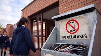 Contenedor metálico lleno de teléfonos con un cartel de prohibición frente al portón de una escuela por el que caminan estudiantes.
