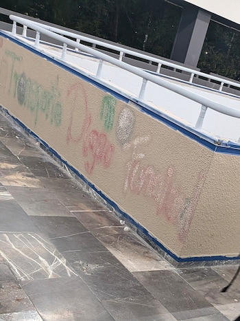 Primer plano de una pared texturizada de color beige con grafitis en rojo, verde y blanco, un pasamanos blanco y suelo de baldosas grises