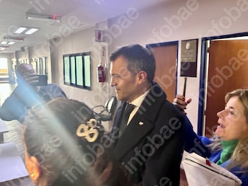 Hombre con traje oscuro, camisa blanca y corbata negra en un pasillo. Una mujer rubia con libreta y bolígrafo junto a él. Otras personas visibles