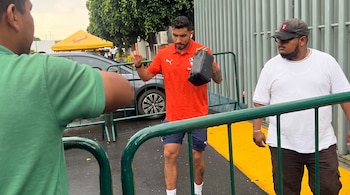 Un hombre con camiseta naranja y bolso de mano camina entre vallas metálicas junto a otras personas