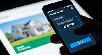 Mano sujetando smartphone con aplicación de transferencia de dinero, monto de $50,000 y botón 'Send'. Fondo desenfocado con imagen de una casa.