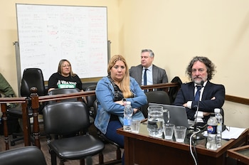 Varias personas sentadas en una sala judicial junto a un escritorio con una computadora, botellas y una pizarra escrita al fondo