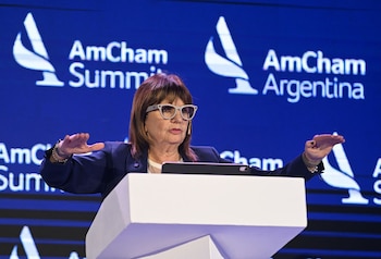Mujer con gafas y blazer oscuro hablando en un podio blanco frente a una pantalla azul con los logos de AmCham Summit y AmCham Argentina