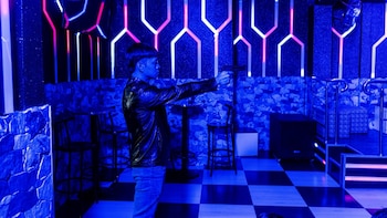 Hombre con chaqueta de cuero negra y jeans azules apunta una pistola en un salón con luces de neón azules y rosadas