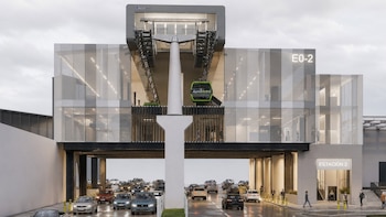 Edificio de cristal y acero etiquetado como Estación 2, sobre una calle con autos y una cabina verde de teleférico en la parte superior
