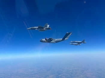Un avión de transporte militar de color gris vuela junto a tres cazas de combate sobre un terreno llano bajo un cielo claro