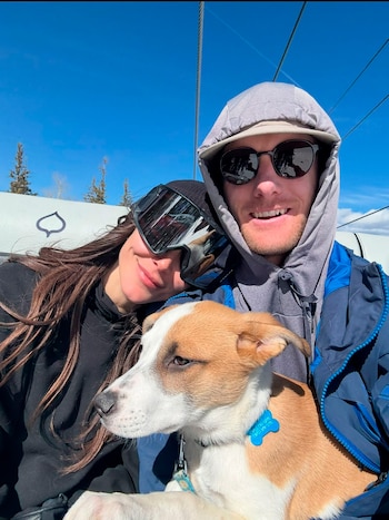 Un hombre y una mujer con gafas de sol posan en un telesilla junto a un perro pequeño blanco y marrón bajo un cielo despejado