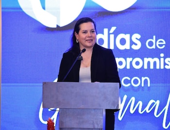 Una mujer con blazer oscuro y top claro de pie frente a un podio con micrófono, sonriendo. Al fondo, una pantalla azul con texto blanco