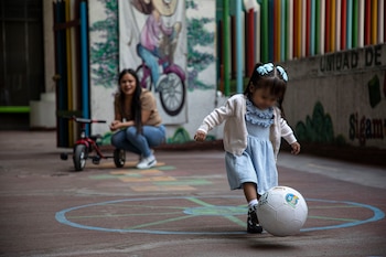 Una niña de dos años con vestido azul claro y cárdigan blanco patea un balón de fútbol blanco en una cancha marrón, mientras su madre observa de fondo
