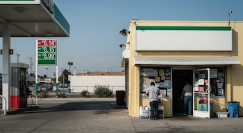 Vista horizontal de una estación de servicio con un letrero de precios de gasolina altos en un barrio urbano de California, junto a un pequeño comercio amarillo con personas.