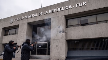 Dos personas con el rostro cubierto disparan armas largas hacia un edificio de concreto con el letrero de la Fiscalía General de la República.