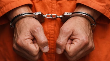Dos manos sujetas por esposas metálicas sobre la parte frontal de un uniforme de color naranja.