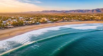 Vista aérea de playa con arena y mar turquesa, olas y surfistas. Un pueblo con palmeras se extiende por la costa, con montañas y cielo dorado de atardecer al fondo.