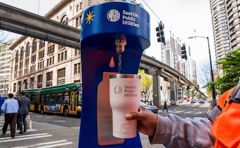 Estación de agua azul de Seattle Public Utilities dispensando agua en una taza blanca. De fondo, una calle urbana con un autobús y edificios