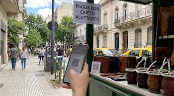 Una mano sostiene un smartphone con una pantalla gris mostrando un código QR para pagos, frente a un puesto callejero con productos de cuero y mates.