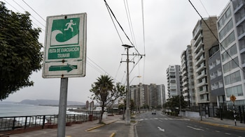 Un cartel verde y blanco de "Ruta de Evacuación Tsunami" con un icono de persona corriendo y una ola, en un poste. Al fondo, la costa, el mar, edificios y una carretera