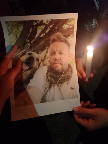 Un hombre sonriente con camiseta blanca y tatuajes acaricia a un perro negro mientras otros perros lo rodean en el campo