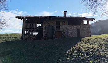 Una casa de piedra y madera con escalera exterior, chimenea y depósito, situada en una colina verde bajo un cielo azul con nubes